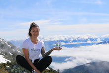 Load image into Gallery viewer, Woman in white PureLife Organics t-shirt sitting cross-legged on a mountain peak, holding a supplement bottle, with snow-capped mountains and clouds in the background